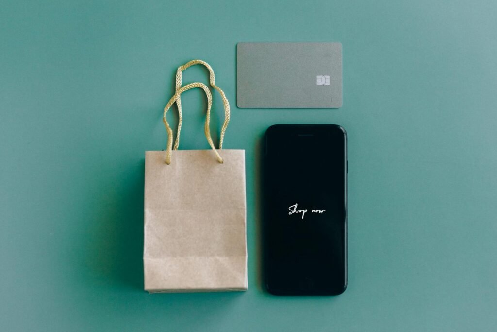 Top view of smartphone with 'Shop now' text, credit card, and paper bag on green background.